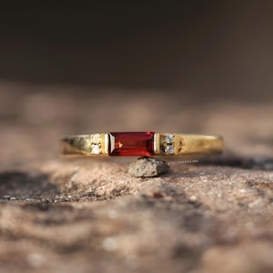 May include: A gold ring with a rectangular, deep red gemstone at the center. The band has small, clear accent stones on either side of the main stone. The ring is set against a blurred, natural background, highlighting the jewelry's design.