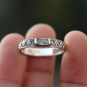 May include: A silver ring featuring a rectangular moss agate stone set in the centre. The band is crafted with an intricate openwork pattern of repeating knot-like motifs. The text RAJSILVERONLINE is engraved on the metal band near the stone.