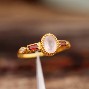 May include: A gold ring featuring a pink oval gemstone with a beaded border. Two red rectangular gemstones are positioned on either side of the centre stone.