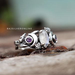 May include: A silver ring featuring a moonstone and amethyst gemstones. The ring has a crescent moon design with floral accents.