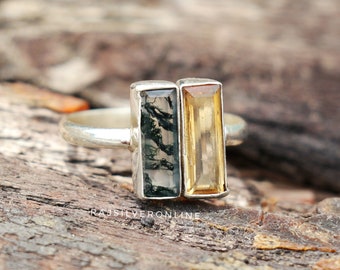 May include: A silver ring featuring two rectangular gemstones. One stone is a moss agate with green inclusions, and the other is a yellow citrine. The ring is set on a wooden surface.
