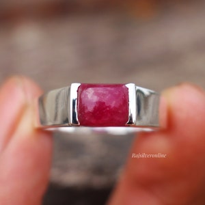 May include: A silver ring with a rectangular, deep red gemstone set in a bezel setting. The ring has a thick band with a polished finish.