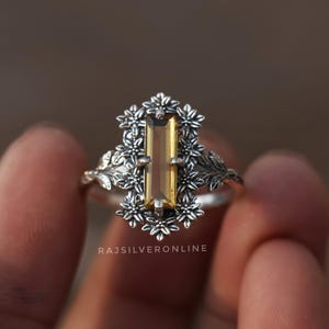 May include: A silver ring with an amber-coloured rectangular gemstone, framed by a floral design. The ring's band has leaf detailing. The text "RAJSILVERONLINE" is visible on the ring.