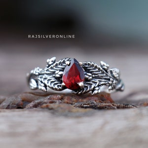 May include: A silver ring with a red gemstone set in a leafy design. The ring is on a wooden surface.