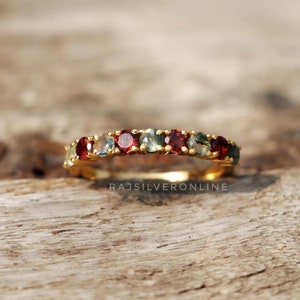 Peut inclure: Une bague en or avec une rangée de pierres précieuses rouges et vertes. La bague est posée sur une surface en bois.