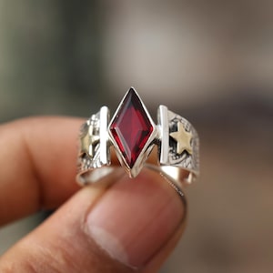 Könnte beinhalten: Ein silberner Ring mit einem großen, roten, rautenförmigen Edelstein in der Mitte. Der Ring hat goldene Sternakzente an den Seiten und ein detailliertes Band. Der Ring wird von einer Hand gehalten.