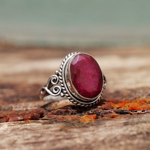 Peut inclure: Une bague en argent avec une grande pierre de rubis de forme ovale. La bague a un design délicat et orné avec une bande torsadée et une bordure décorative autour de la pierre.