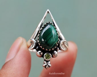 May include: A silver ring featuring a large, teardrop-shaped, dark green malachite stone. The ring has a unique design with a triangular top, crescent moon accents, and a small, round, light green stone. The text "rajsilveronline" is visible.