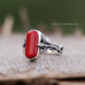Puede incluir: Un anillo de plata con una piedra de coral rojo y un diseño de hojas. El anillo está sobre una superficie de madera. RAJSILVERONLINE está impreso en el fondo.