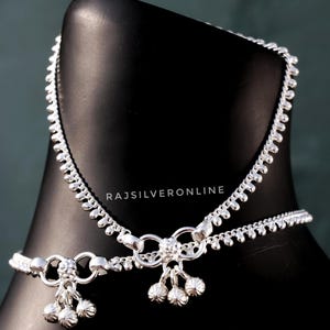 May include: A pair of silver anklets displayed on a black mannequin. The anklets feature a delicate chain design with small, round accents and dangling bell charms. The text "RAJSILVERONLINE" is visible.