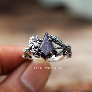 Peut inclure: Une bague en argent avec une pierre précieuse violette foncée entourée d'un motif floral. La bague présente une délicate fleur et des feuilles.