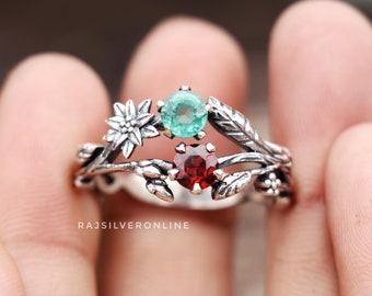 May include: A silver ring with a delicate floral design featuring a green and red gemstone. The ring is set on a finger.