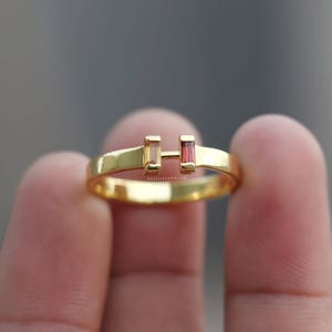 May include: A gold ring featuring an open-gap design with two baguette-cut gemstones, one yellow and one red, set vertically at the ends. The band is polished gold, and the text RAJSILVERONLINE is engraved on the inner surface of the ring band.