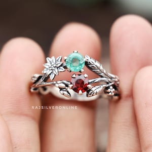 Könnte beinhalten: Ein silberner Ring mit einem zarten floralen Design, das mit einem grünen und roten Edelstein verziert ist. Der Ring sitzt an einem Finger.