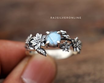 May include: A silver ring with a blue gemstone surrounded by delicate flower designs. The ring is set on a wooden surface.