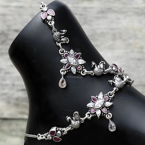 Op de afbeelding: Zilveren enkelband met bloemen- en pauwontwerpen. De enkelband heeft heldere en bordeauxkleurige edelstenen. Het ontwerp bevat druppelvormige bedels en wordt getoond op een zwarte voetvorm.