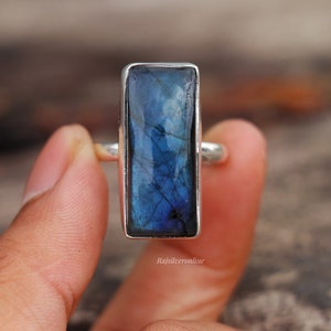 May include: A rectangular labradorite ring with a silver band. The gemstone displays a vibrant blue color with internal veining. The ring is held by a person's fingers, showcasing the polished stone and the silver setting.