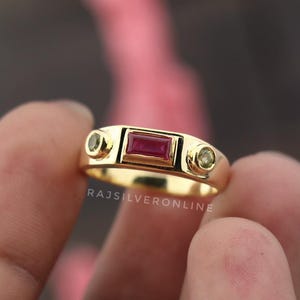 Pode incluir: Um anel dourado com uma pedra retangular cor de rubi no centro e duas pedras redondas menores, tingidas de verde, em ambos os lados. O anel tem um acabamento liso e polido. O texto "RAJSILVERONLINE" é visível.