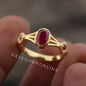 May include: A gold ring with a central oval-cut, deep red gemstone. The ring has a detailed design with smaller red stones on either side. The text "RAJSILVERONLINE" is visible on the ring.