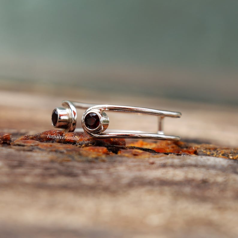 May include: A silver ring with two red gemstones set in the band. The ring is designed with two parallel bands that are connected by a small loop.