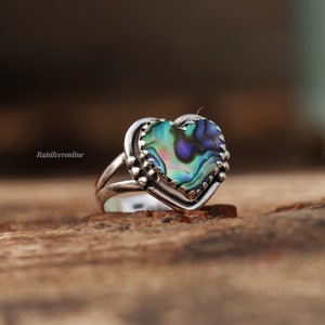 May include: A silver ring featuring a heart-shaped abalone shell inlay. The ring has a textured edge and a delicate design.