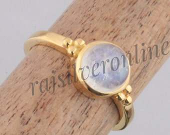 Puede incluir: Un anillo de tono dorado con un cabochón de piedra lunar redondo y translúcido engastado en un bisel liso. La banda incluye pequeños grupos decorativos de tres diminutas esferas doradas a cada lado de la piedra central. El texto rajsilveronline es visible.