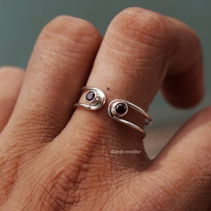 May include: A silver ring with two small, dark red gemstones. The ring is open and adjustable.