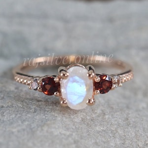 Könnte beinhalten: Ein Ring aus Roségold mit einem großen ovalen Mondstein und zwei kleineren runden Granatsteinen auf jeder Seite. Die Band ist mit kleinen weißen Steinen verziert.