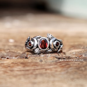 May include: A silver ring with a red gemstone and floral designs. The ring features three crescent moons and three red gemstones.