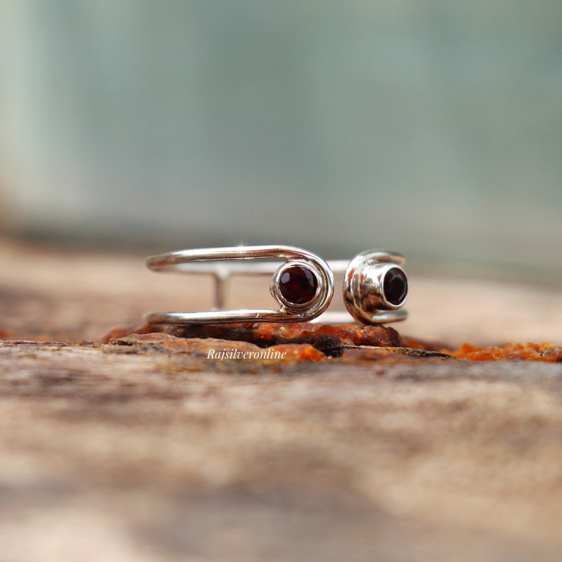 May include: A silver ring with two red gemstones. The ring is made of two bands that are connected by a small loop. The ring is on a wooden surface.