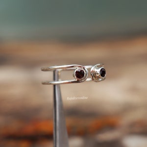 May include: A silver ring with two red gemstones. The ring is designed with two bands that are connected by a small loop. The ring is on a display stand.
