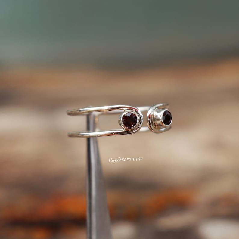 May include: A silver ring with two red gemstones. The ring is designed with two bands that are connected by a small loop. The ring is on a display stand.