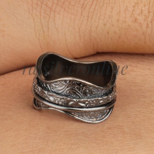 May include: A dark silver-toned ring with an intricate floral pattern. The ring has a spinning band and a curved design. The ring is shown on a person's skin.