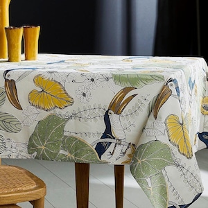 Op de afbeelding: Wit tafelkleed met een tropische print met toekans, bladeren en bloemen. Het tafelkleed ligt op een tafel met een houten stoel.
