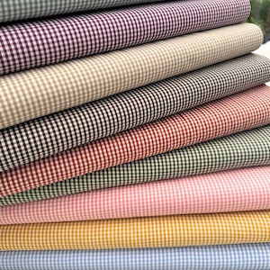 Può includere: Una pila di rotoli di tessuto piegati con un motivo a quadretti vichy di piccole dimensioni in vari colori, tra cui azzurro, viola, beige, marrone, rosso, verde scuro, rosa, giallo e nero, disposti in un ordine orizzontale sovrapposto.