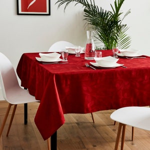 Peut inclure: Une nappe rouge recouvre une table en bois dressée pour un repas. Des assiettes, des verres et des couverts blancs sont disposés sur la table. Une carafe en verre transparent et une plante verte sont en arrière-plan.