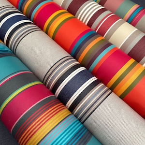 Pode incluir: V&aacute;rios rolos de tecido dispostos na diagonal, cada um apresentando padr&otilde;es distintos de riscas verticais em paletas de cores vibrantes, incluindo laranja, azul-petr&oacute;leo, magenta, azul-marinho, cinzento e creme. As superf&iacute;cies t&ecirc;xteis mostram uma textura tecida.