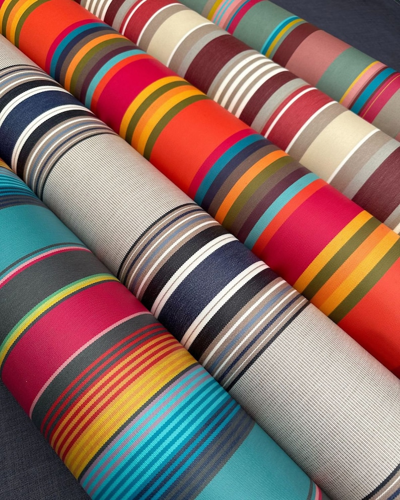 Pode incluir: V&aacute;rios rolos de tecido dispostos na diagonal, cada um apresentando padr&otilde;es distintos de riscas verticais em paletas de cores vibrantes, incluindo laranja, azul-petr&oacute;leo, magenta, azul-marinho, cinzento e creme. As superf&iacute;cies t&ecirc;xteis mostram uma textura tecida.