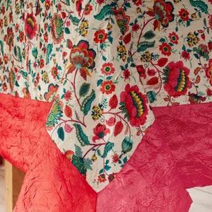 Puede incluir: Un mantel blanco con un patrón floral en tonos de rosa, verde y amarillo. El mantel está doblado por la mitad y los bordes están recortados con una tela rosa brillante.