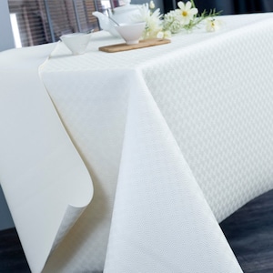 May include: White tablecloth with a subtle embossed pattern. The tablecloth is laid on a table with a wooden cutting board, two white bowls, and a white teapot.