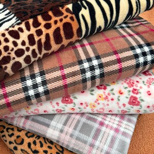 Könnte beinhalten: Ein Stapel gefalteter Fleece-Stoffstücke mit verschiedenen Mustern, darunter Leopardenmuster, Zebrastreifen, ein beigefarbenes Karomuster mit schwarzen und weißen Linien und dünnen roten Akzenten, ein weißer Blumendruck und ein graues Karomuster.