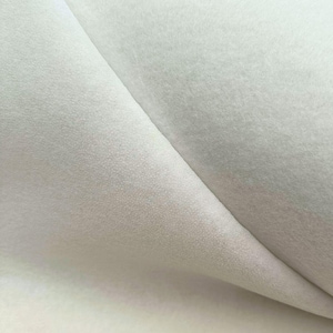 Può includere: Primo piano di un tessuto bianco sporco e morbido con una texture vellutata. Il tessuto è piegato, rivelando ombre sottili e variazioni di tono. Il materiale appare liscio e soffice, adatto a vari progetti artigianali o tessili.