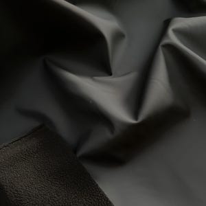 May include: Close-up of black fabric with a smooth, shiny surface. The fabric is draped and folded, creating shadows and highlights. A section of textured, dark material is visible in the lower left corner, contrasting with the smooth fabric.