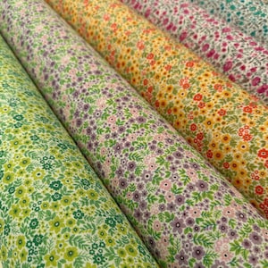 Könnte beinhalten: Mehrere Stoffrollen mit floralen Mustern in verschiedenen Farben. Die Designs umfassen kleine Blumen und Blätter in Grün-, Gelb-, Rosa- und Lilatönen. Die Rollen sind diagonal angeordnet und zeigen die verschiedenen Muster.