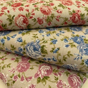 Puede incluir: Tres capas plegadas de tela tejida con un estampado floral sobre un fondo beige claro. La capa superior muestra rosas rojas, la capa intermedia rosas azules y la capa inferior rosas de color rosa, todas con hojas y tallos verdes.