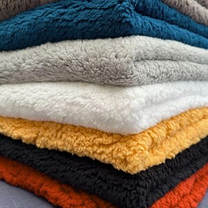 Puede incluir: Una pila de siete mantas de felpa en varios colores, incluyendo gris, azul verdoso, gris claro, blanco, amarillo, negro y rojo. Las mantas están dobladas y apiladas una encima de la otra.