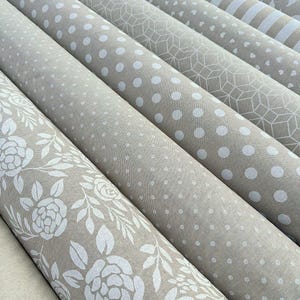 Peut inclure: Plusieurs rouleaux de tissu avec divers motifs. Les motifs incluent des motifs floraux blancs, des pois, des formes g&eacute;om&eacute;triques et des rayures, le tout sur un fond beige neutre. Le tissu semble &ecirc;tre du coton ou du lin.