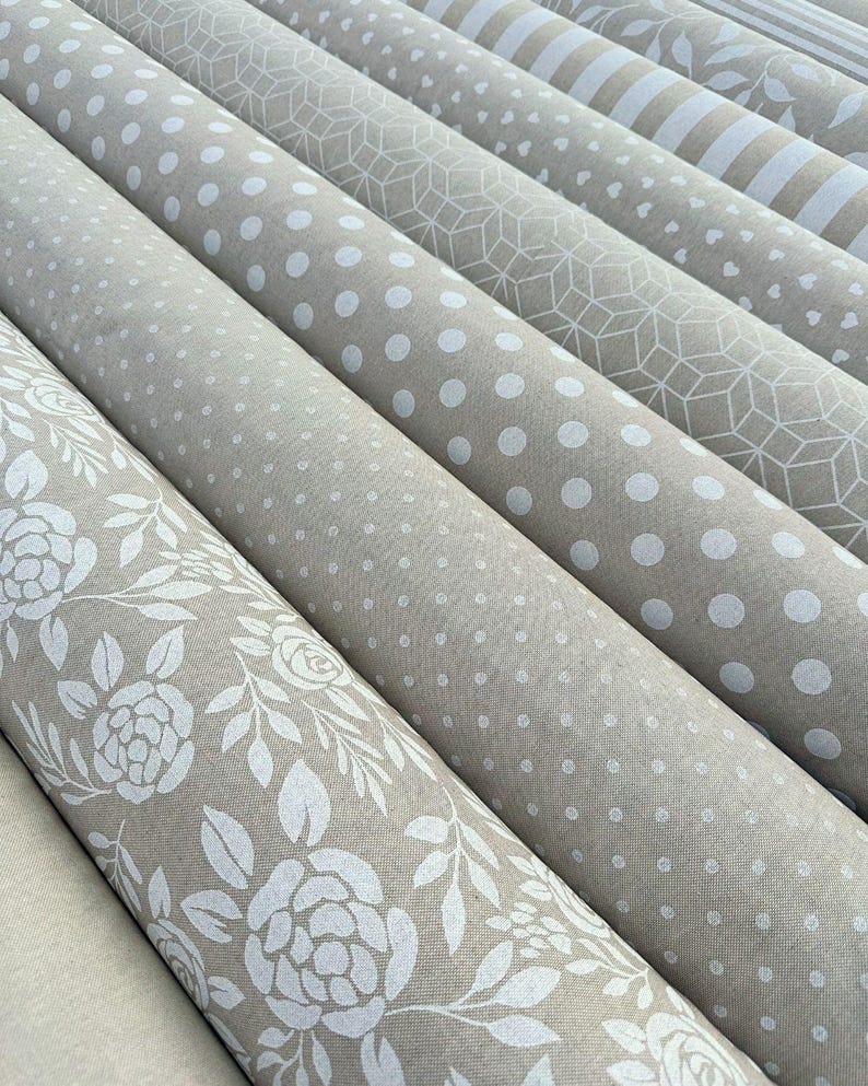 Peut inclure: Plusieurs rouleaux de tissu avec divers motifs. Les motifs incluent des motifs floraux blancs, des pois, des formes g&eacute;om&eacute;triques et des rayures, le tout sur un fond beige neutre. Le tissu semble &ecirc;tre du coton ou du lin.