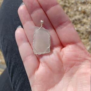 Puede incluir: Un colgante de vidrio transparente envuelto en alambre plateado. El colgante tiene forma rectangular y una superficie lisa y pulida.