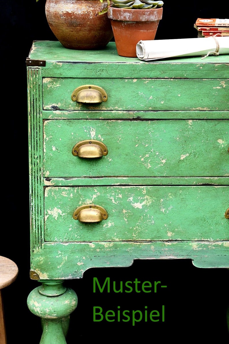 K&ouml;nnte beinhalten: Eine gr&uuml;n lackierte Kommode aus Holz mit vier Schubladen und Messinggriffen. Die Kommode hat eine abgenutzte Oberfl&auml;che und steht auf einem schwarzen Hintergrund. Der Text "Muster-Beispiel" ist in Gr&uuml;n unter der Kommode geschrieben.